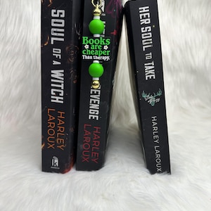 May include: Three hardcover books with black spines and white text. A green and gold beaded bookmark is between two books. The bookmark has a black rectangle with the text "Books are cheaper than therapy."