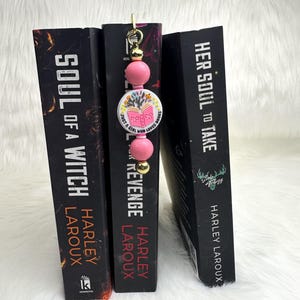 Puede incluir: Un marcapáginas de cuentas rosas con un dije que dice "Just a girl who loves books" se coloca entre tres libros. Los títulos de los libros son "Soul of a Witch", "Revenge" y "Her Soul to Take" de Harley Laroux.