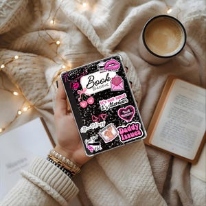 May include: A black notebook-style phone case with various pink and white stickers, including lips, cherries, and the words "Book Baddie" and "Daddy Issues." The case is held in a hand, with a cup of coffee and a book in the background.