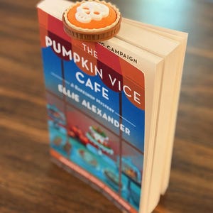 May include: A book titled The Pumpkin Vice Cafe by Ellie Alexander with a round, orange, pie-shaped bookmark featuring a white skull on top. The bookmark is clipped to the top edge of the book pages, which are white and rectangular in shape.