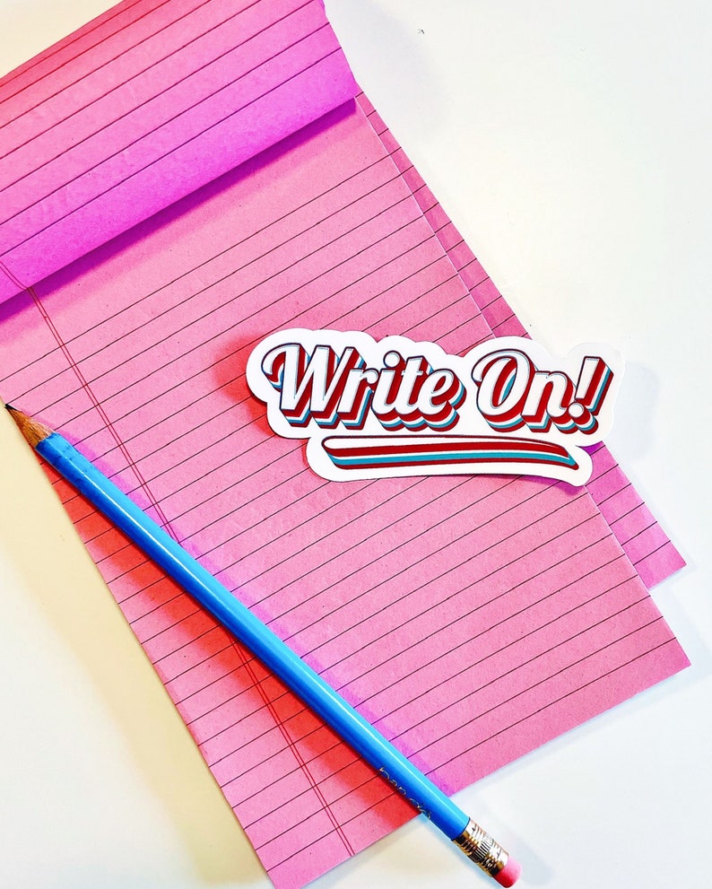 以下が含まれることがあります： ピンクの罫線入りノートに青い鉛筆と、「Write On!」と赤、青、白の文字で書かれた白いステッカーが貼ってあります。