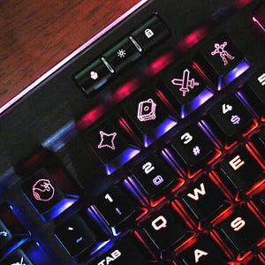 May include: Close-up of a black gaming keyboard with illuminated keys. The keys are backlit with red and blue lights. The keyboard features various symbols and letters, including numbers, letters, and special characters. The keyboard is on a wooden surface.