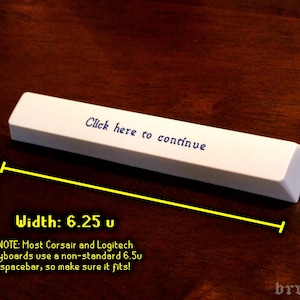 K&ouml;nnte beinhalten: Eine wei&szlig;e Tastatur-Leertaste mit dem Text "Click here to continue". Die Leertaste ist 15,88 cm breit. Das Bild befindet sich auf einer dunkelbraunen Oberfl&auml;che.