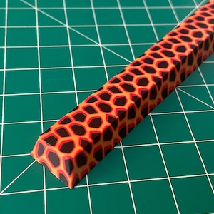 Może przedstawiać: Prostokątny przedmiot z wzorem plastra miodu w odcieniach pomarańczu, czerwieni i czerni. Obiekt ma fazowaną krawędź i jest umieszczony na zielonym tle w kratkę. Wzór przypomina teksturę lawy lub ognia.