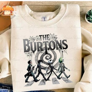 May include: A beige sweatshirt with a graphic print of characters from The Nightmare Before Christmas and Beetlejuice walking across a crosswalk. The text "The Burtons" is printed above the characters.