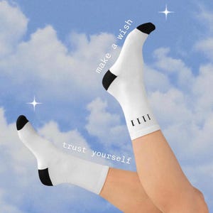 May include: White socks with black trim and the text "make a wish" and "trust yourself" on them. The socks are worn by a person's legs against a blue sky background with white clouds.
