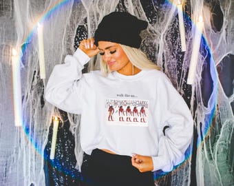 Pode incluir: Camisola branca cropped com o texto "walk like an..." e um gráfico de cinco figuras em roupas vermelhas e brancas. A camisola tem mangas compridas e um caimento descontraído.