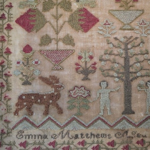 Op de afbeelding: Een gedetailleerd antiek borduurwerk met een beige achtergrond. Het borduurwerk toont een hert, bomen, bloemen en geometrische vormen in rood, groen en bruin. De naam "Emma Matthews" is onderaan geborduurd.