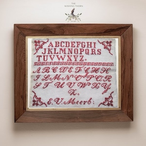 May include: A framed sampler with the alphabet embroidered in red thread on white fabric. The sampler is signed "E.U. Meerb."