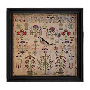 Può includere: Un ricamo incorniciato con un uccello, fiori e alberi ricamati con fili colorati su un tessuto color crema. Il ricamo è datato 1873 e firmato "Anne Davies".