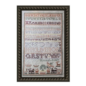 Könnte beinhalten: Ein gerahmter Kreuzstich-Sampler mit weißem Hintergrund und bunter Stickerei. Der Sampler enthält das Alphabet, Zahlen und eine kleine Szene mit einem Vogel, einem Baum und einem Reh. Der Text "Hannah Rider" ist unten gestickt.