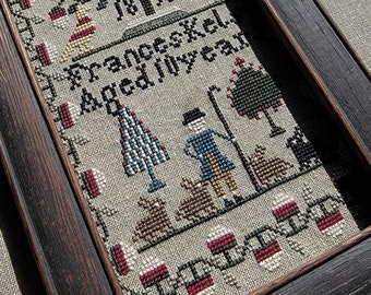 Pode incluir: Amostra de ponto cruz emoldurada com uma cena pastoral com uma figura, ovelhas e árvores. A amostra inclui o texto "Frances Kel. Aged 10 years" e a data "1815". A moldura é de madeira escura.