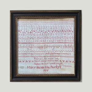 Könnte beinhalten: Ein gerahmtes antikes Stickmuster mit roter Kreuzstichstickerei auf cremefarbenem Stoff. Das Muster enthält das Alphabet, Zahlen und einen Vers, mit dem Namen "Mary Heneritta Purdy" und dem Datum 1818. Der Rahmen ist dunkelbraun.