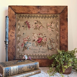 May include: A framed antique needlepoint sampler featuring floral designs in shades of pink, red, and blue on a tan background. The sampler is dated September 8th, 1847, and is in a rustic wooden frame. A candle and books are in the foreground.