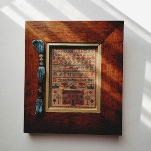 May include: A framed cross-stitch sampler with a brown wooden frame. The sampler features a house with a red door and a garden with trees. The sampler is dated 1888 and signed "Fiona Rutherford".