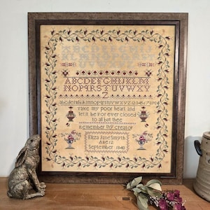 以下が含まれることがあります： クリーム色の背景と、えんじ色、緑色、茶色の花柄の縁取りが施された、額装されたクロスステッチのサンプル。サンプルにはアルファベット、数字、そして「私の貧しい心を持ち、あなた以外の人には永遠に閉じられたままにしてください。あなたの創造者を忘れないでください。」という詩が書かれています。サンプルには「Eliza Jane Smyth、Ase12、1840年9月」と署名されています。