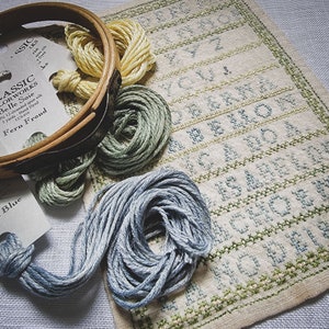 May include: A close-up of a cross-stitch embroidery project with the alphabet stitched in green and blue thread. The project is framed in a wooden embroidery hoop with skeins of blue, green, and yellow embroidery floss.