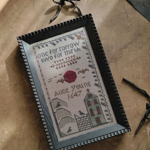 Puede incluir: Un sampler de punto de cruz enmarcado con un marco negro y un fondo blanco. El sampler presenta un sol rojo, dos casas y el texto "One for sorrow, two for mirth, Alice Youngs 1647".