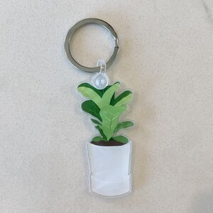 May include: A white potted plant keychain with a green leafy plant. The keychain has a silver metal ring.