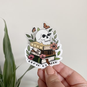 May include: A white skull with a stack of books underneath it. The books are decorated with flowers and butterflies. The text "Death by TBR" is written below the books.
