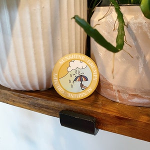 May include: A yellow and blue round sticker with the text "Sunshine Grumpy Sunshine Grumpy" and a cartoon illustration of a cloud with raindrops and an umbrella.