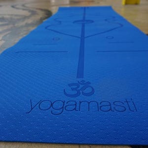 May include: A blue yoga mat with a white Om symbol and the text "yogamasti" printed on it. The mat has a textured surface and a line down the centre.
