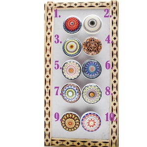 Può includere: Un'esposizione di dieci pomelli decorativi per armadi con disegni mandala colorati e intricati. I pomelli sono disposti su uno sfondo bianco all'interno di una cornice di legno. Ogni pomello presenta un motivo unico con vari colori.