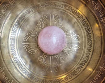 Peut inclure: Une sphère en quartz rose est placée au centre d'un bol chantant en laiton. Le bol présente des motifs gravés complexes et des inscriptions sur le bord. La sphère est de couleur rose pâle.