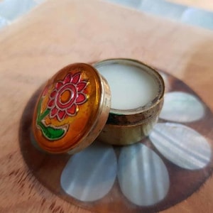 Puede incluir: Un pequeño recipiente metálico vintage abierto, con un diseño floral en la tapa. La tapa es naranja con una flor roja y amarilla y hojas verdes. El recipiente está lleno de una sustancia blanca.