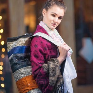 May include: A woman wearing a pink and black patterned shawl carries a colourful yoga mat bag with a blue strap. The bag is made of fabric with a variety of textures and patterns.