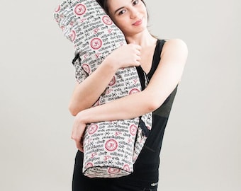 May include: A white yoga mat bag with black Sanskrit text and red Om symbols. The bag is being held by a person wearing a black tank top and black skirt.