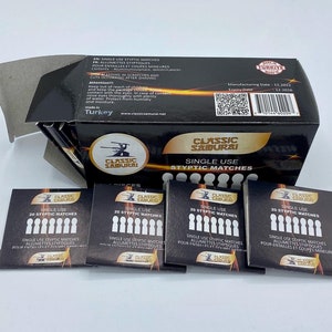 Peut inclure: Une boîte de 20 allumettes styptiques à usage unique, étiquetée "Classic Samurai". Les allumettes sont blanches et sont présentées dans une petite boîte noire avec un logo de samouraï doré. La boîte comprend également le texte "Allumettes styptiques à usage unique" et "Pour entailles et coupures mineures".