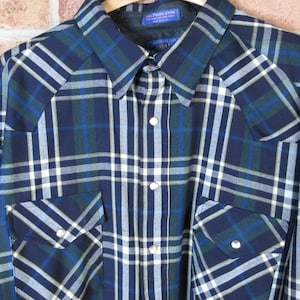 May include: A long-sleeved plaid shirt in navy blue, white, and green, hanging on a wooden hanger. The shirt features two chest pockets with button closures and a classic collar. The plaid pattern is a mix of vertical and horizontal lines.