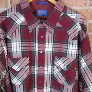 Puede incluir: Una camisa de cuadros de manga larga en rojo, blanco y gris, con dos bolsillos delanteros y cuello. La camisa est&aacute; colgada de una percha de madera. La camisa tiene una botonadura frontal y una etiqueta "Pendleton".