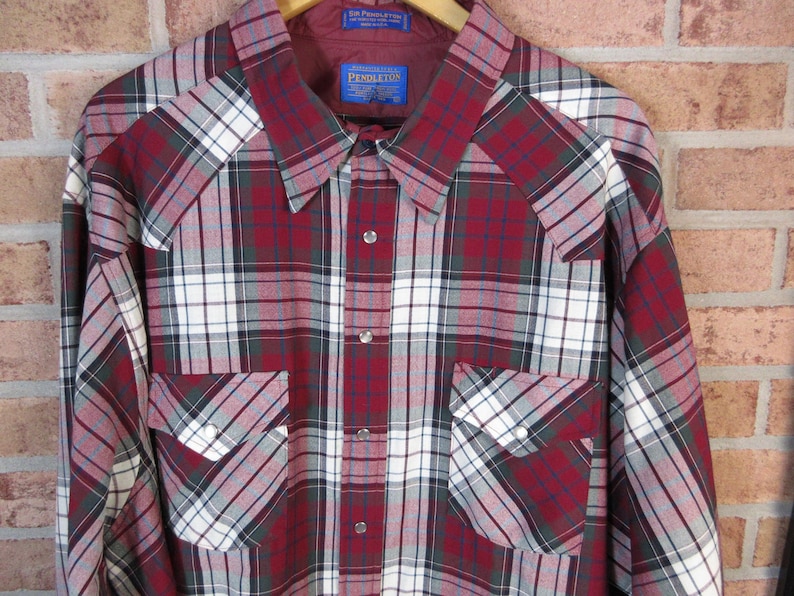 Puede incluir: Una camisa de cuadros de manga larga en rojo, blanco y gris, con dos bolsillos delanteros y cuello. La camisa est&aacute; colgada de una percha de madera. La camisa tiene una botonadura frontal y una etiqueta "Pendleton".