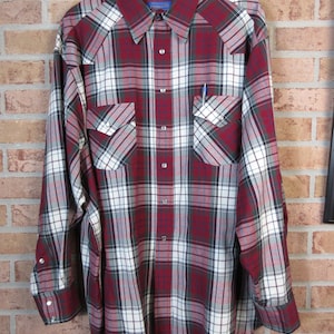 Puede incluir: Una camisa de cuadros de manga larga en tonos rojos, blancos y grises, con botones y dos bolsillos en el pecho. La camisa est&aacute; colgada en una percha de madera contra una pared de ladrillos. La marca "Pendleton" es visible.