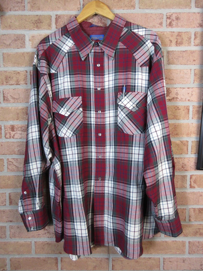 Puede incluir: Una camisa de cuadros de manga larga en tonos rojos, blancos y grises, con botones y dos bolsillos en el pecho. La camisa est&aacute; colgada en una percha de madera contra una pared de ladrillos. La marca "Pendleton" es visible.