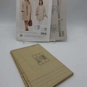 May include: A sewing pattern from Burda Style, featuring a coat and jacket design. The pattern packet includes images of the finished garments and the number 6802. A stack of folded pattern pieces is in the foreground.
