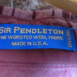 Puede incluir: Primer plano de una etiqueta azul con texto dorado: "SIR PENDLETON FINE WORSTED WOOL FABRIC MADE IN U.S.A." La etiqueta est&aacute; cosida sobre una tela burdeos con un dise&ntilde;o a rayas.