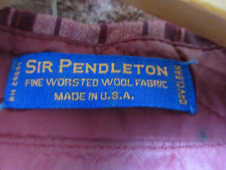 Puede incluir: Primer plano de una etiqueta azul con texto dorado: "SIR PENDLETON FINE WORSTED WOOL FABRIC MADE IN U.S.A." La etiqueta est&aacute; cosida sobre una tela burdeos con un dise&ntilde;o a rayas.