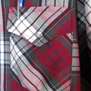 Puede incluir: Primer plano de una camisa a cuadros roja, blanca y azul con un bolsillo. El bolsillo tiene un cierre de bot&oacute;n a presi&oacute;n plateado y un bol&iacute;grafo azul en el bolsillo. La camisa tiene un estilo occidental cl&aacute;sico.