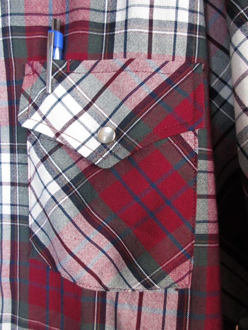 Puede incluir: Primer plano de una camisa a cuadros roja, blanca y azul con un bolsillo. El bolsillo tiene un cierre de bot&oacute;n a presi&oacute;n plateado y un bol&iacute;grafo azul en el bolsillo. La camisa tiene un estilo occidental cl&aacute;sico.