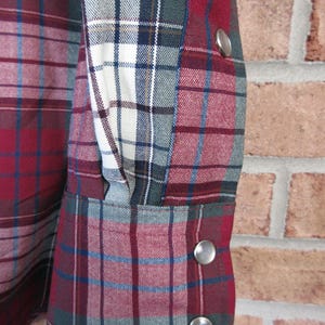 Puede incluir: Primer plano de una manga de camisa a cuadros. La camisa presenta un patr&oacute;n de cuadros rojo, blanco, azul y verde. La manga tiene cierres de presi&oacute;n plateados. La tela parece ser un material tejido.