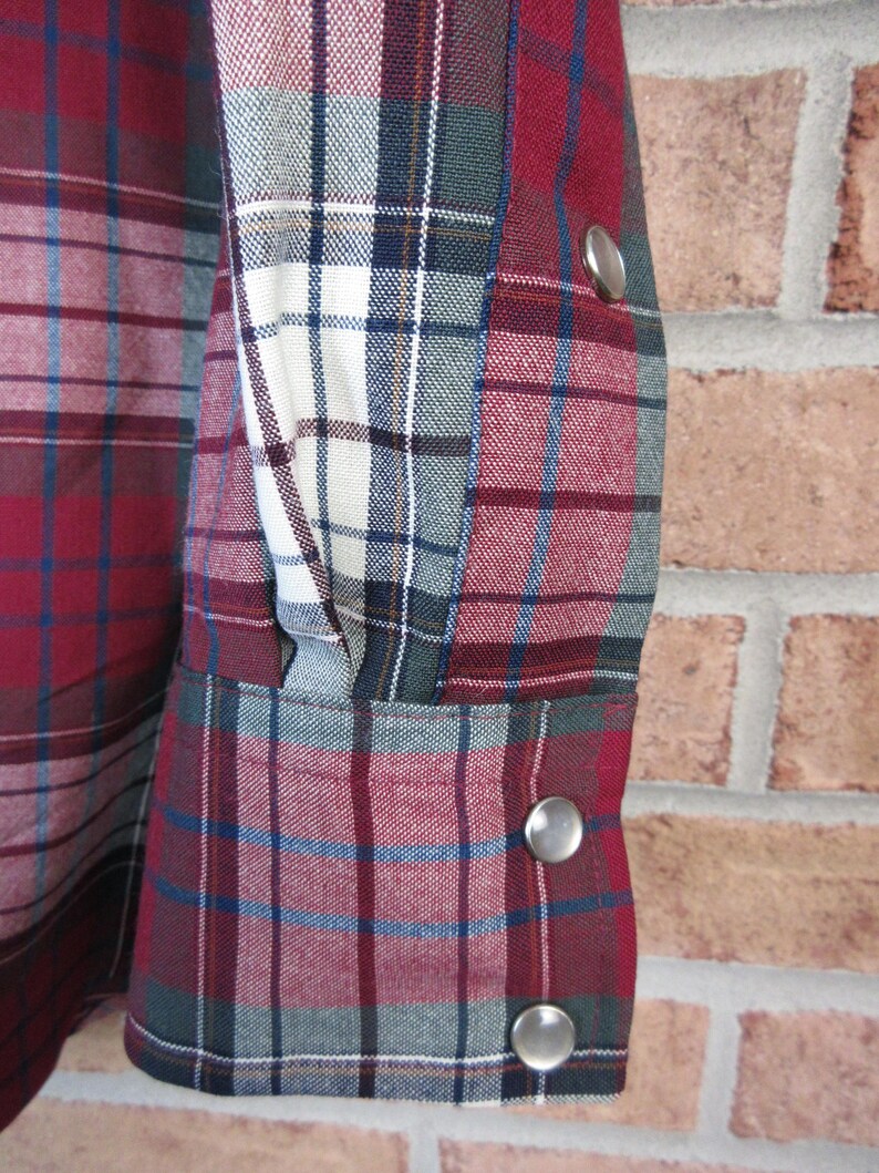 Puede incluir: Primer plano de una manga de camisa a cuadros. La camisa presenta un patr&oacute;n de cuadros rojo, blanco, azul y verde. La manga tiene cierres de presi&oacute;n plateados. La tela parece ser un material tejido.