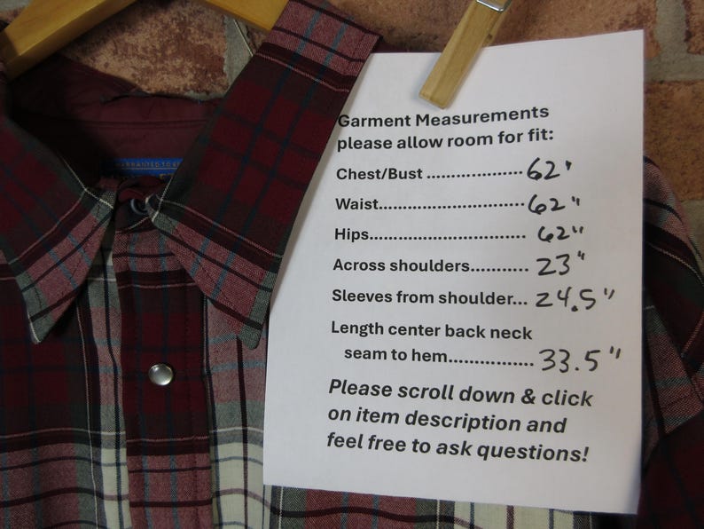 Puede incluir: Una camisa abotonada a cuadros burdeos, grises y blancos con cuello y cierre a presi&oacute;n. Las medidas de la prenda se enumeran en un papel adjunto con una pinza. Las medidas incluyen pecho, cintura, caderas, hombros, largo de manga y espalda.