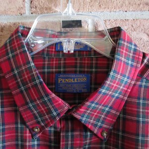 May include: A red, black, and green plaid button-down shirt with a collar and buttons. The shirt has a Pendleton label. The shirt is hanging on a clear plastic hanger against a brick wall.