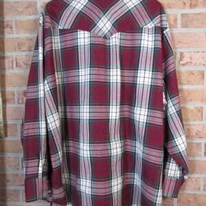 Puede incluir: Camisa de cuadros de manga larga en un patr&oacute;n rojo, blanco y verde oscuro. La camisa est&aacute; colgada de una percha de madera contra una pared de ladrillo. La camisa tiene pu&ntilde;os abotonados y un cuello cl&aacute;sico.