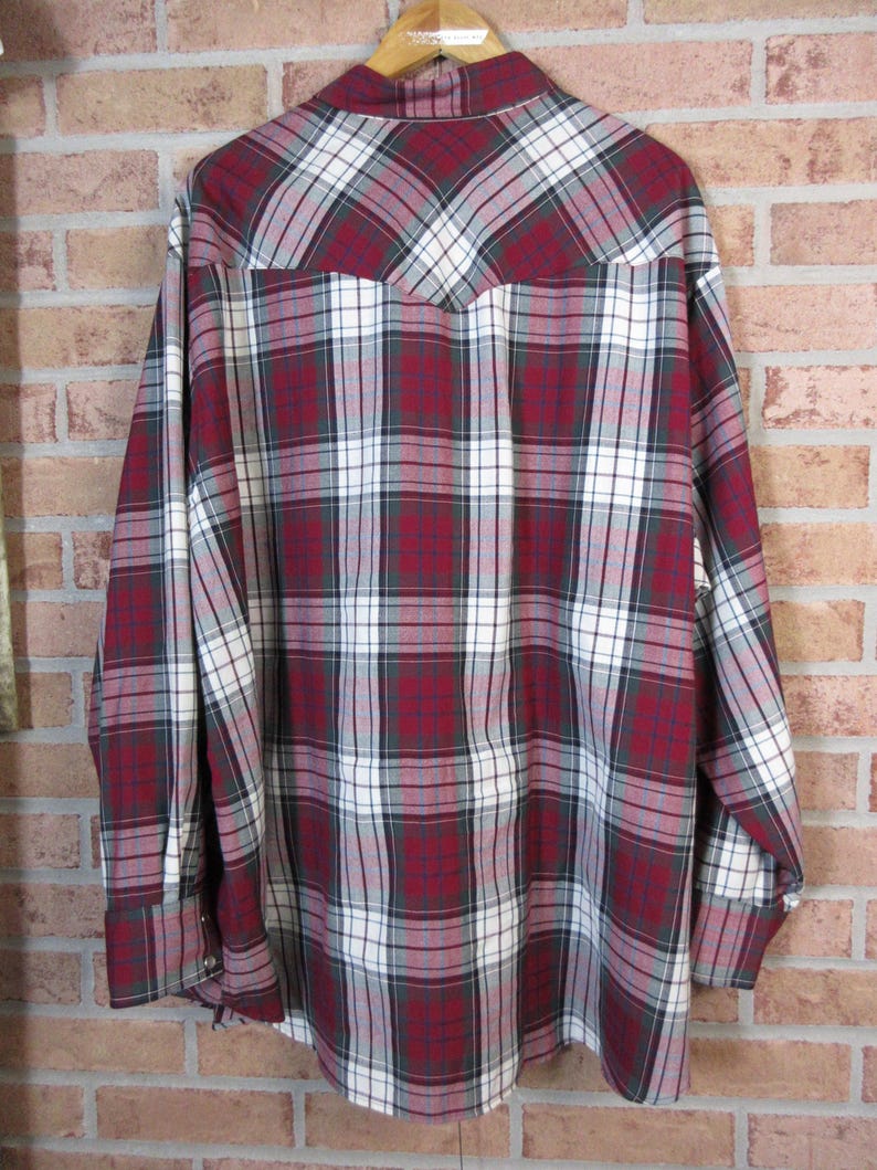 Puede incluir: Camisa de cuadros de manga larga en un patr&oacute;n rojo, blanco y verde oscuro. La camisa est&aacute; colgada de una percha de madera contra una pared de ladrillo. La camisa tiene pu&ntilde;os abotonados y un cuello cl&aacute;sico.