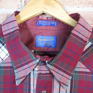 Puede incluir: Primer plano de una camisa a cuadros rojos, azules y blancos con cuello y forro burdeos. La camisa tiene dos etiquetas azules con texto blanco. La camisa est&aacute; colgada de una percha de madera.