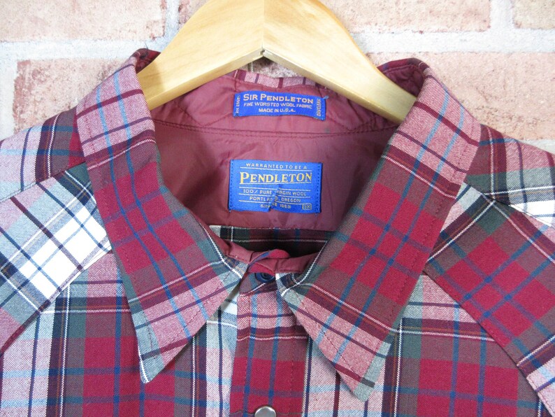 Puede incluir: Primer plano de una camisa a cuadros rojos, azules y blancos con cuello y forro burdeos. La camisa tiene dos etiquetas azules con texto blanco. La camisa est&aacute; colgada de una percha de madera.